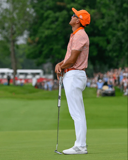 Golfer in an orange hat and striped shirt holding a putter, looking up thoughtfully on a green golf course with a crowd in the background.