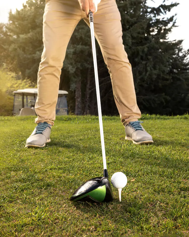 Person standing on a golf course, preparing to hit a golf ball on a tee with a driver club. Trees and a golf cart are visible in the background—a perfect setting to practice and improve putting for your overall game.