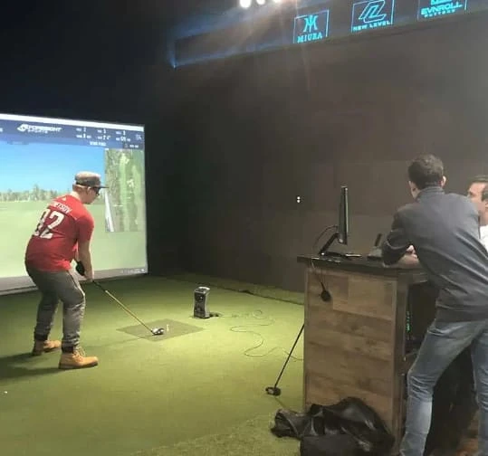 A person swings a golf club at an indoor golf simulator while two others watch near a computer on a wooden desk.