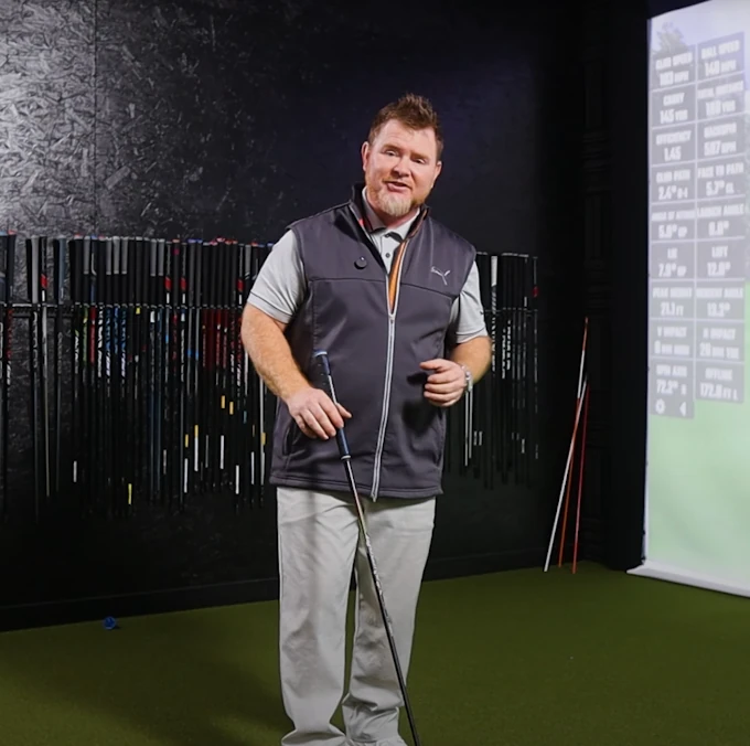 A man stands indoors on artificial turf holding a golf club, with golf clubs on a rack behind him and a projection screen displaying data to his right.