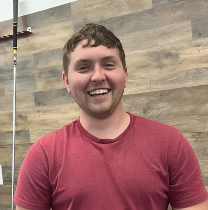 A young man in a red t-shirt smiles in front of a wood-paneled wall, with part of a fishing rod visible in the background.