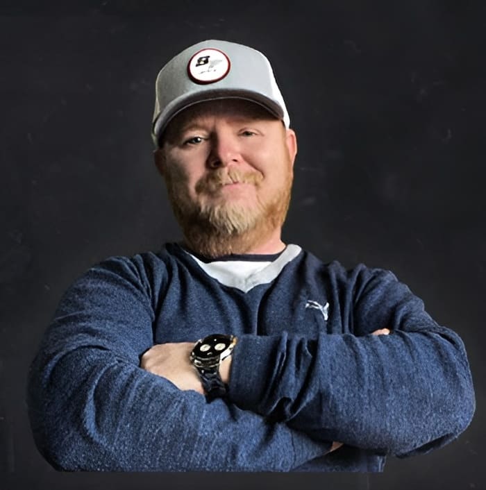 A man with a blond beard wearing a gray cap, blue pullover, and smartwatch stands with his arms crossed against a plain dark background.