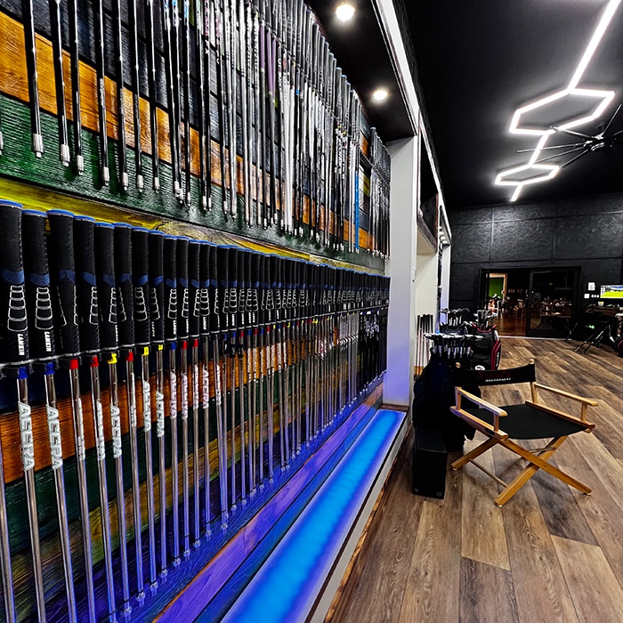 Rows of golf clubs are neatly displayed on a wall in a modern, well-lit indoor space with wood flooring and lounge seating.