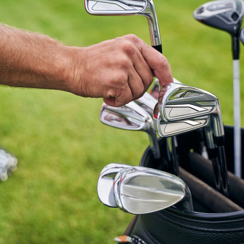 A person selects a golf club from a golf bag containing several irons, with a grassy area in the background.