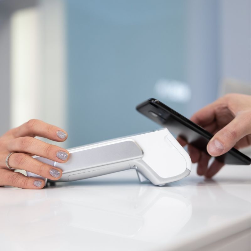 A person holds a smartphone near a card reader to make a contactless payment, while another person holds the device on a white countertop.